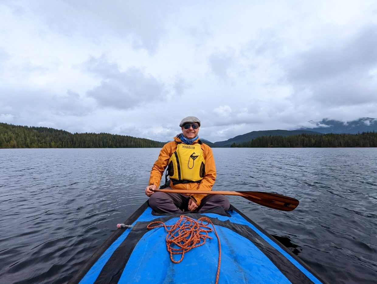 A man sits in a canoe in the middle of a lake. The canoe has a tarp on it and the sky is cloudy.