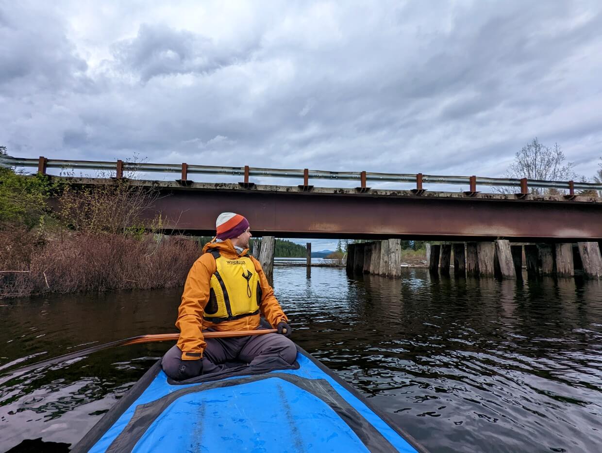 Man sitting in a canoe looking back at Brewster bridge. The sky is overcast.