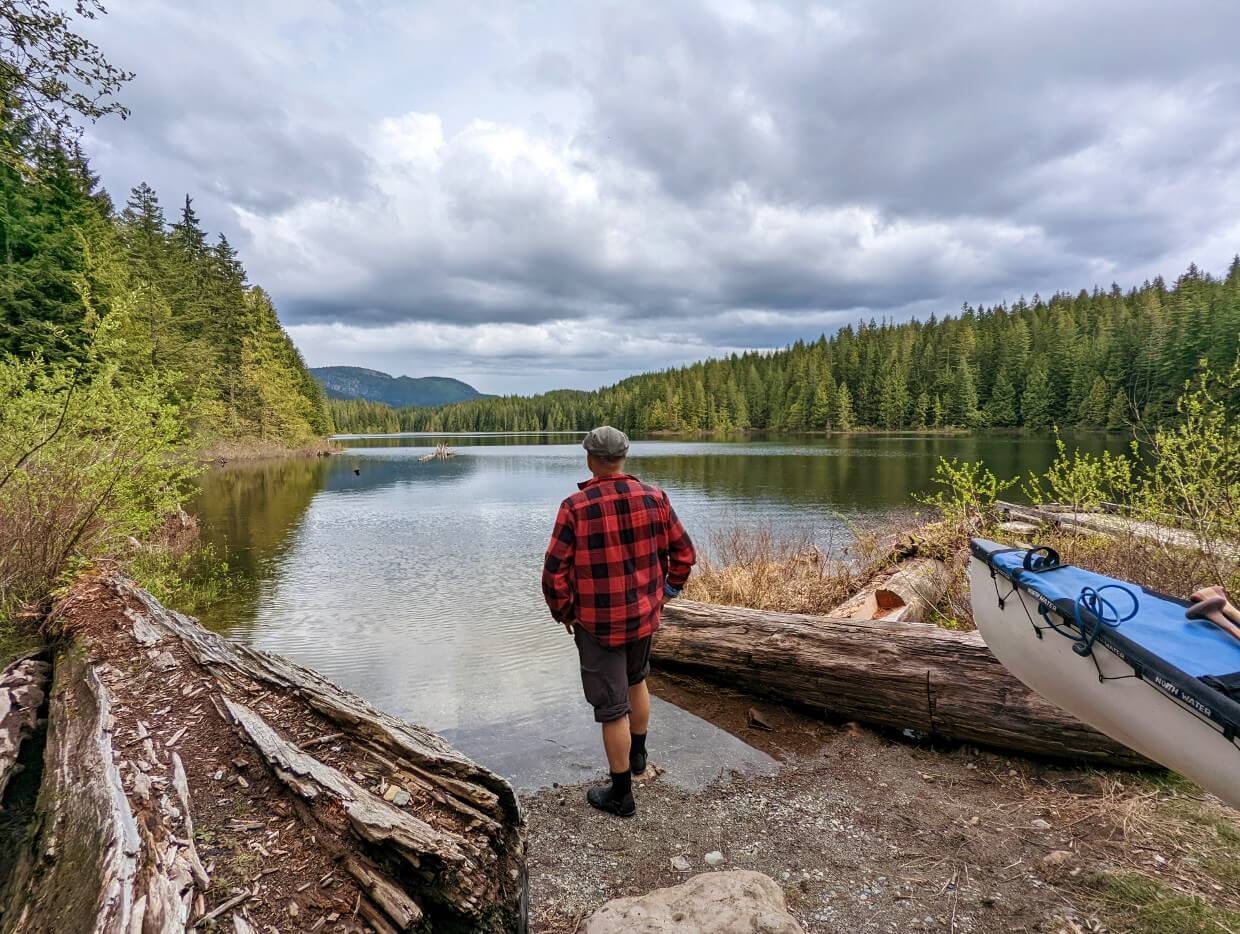 Man standing on the edge of Twin Lakes looking out at the view. To the right sits a canoe and and old fallen tree. The lake is surrounded by lush green forests.