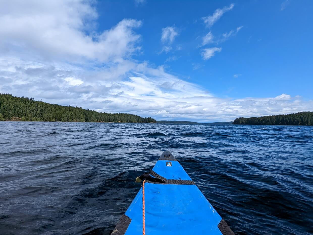Tip of a canoe on Lower Campbell Lake. The water is very choppy. In the distance forest hugs the shoreline and the sky is blue with puffy white clouds.