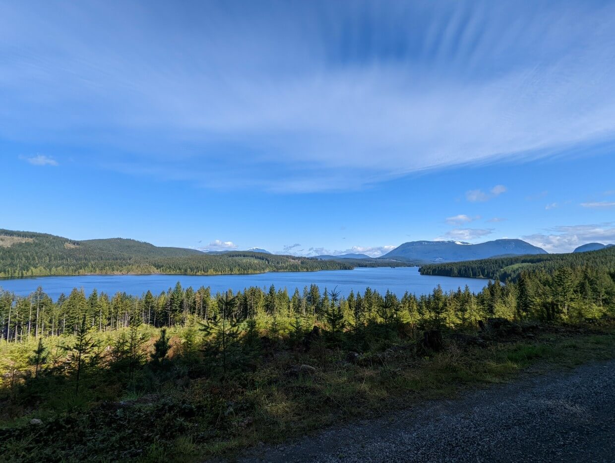 View of Mohun Lake from Morton Lake Road. In between the road and the lake is a stretch of forest. On the other side of the lake is more forest and mountains in the distance under a blue sky.