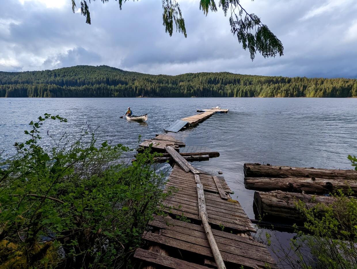 An abandoned and disheveled dock reaching out onto Mohun Lake. A man paddling in a canoe is approaching the dock in the water.