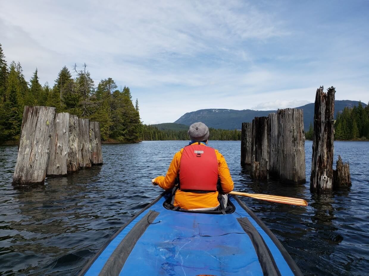 Man paddling through the remains of an old railway trestle. Trees hug the lake and in the distance mountain can be seen.