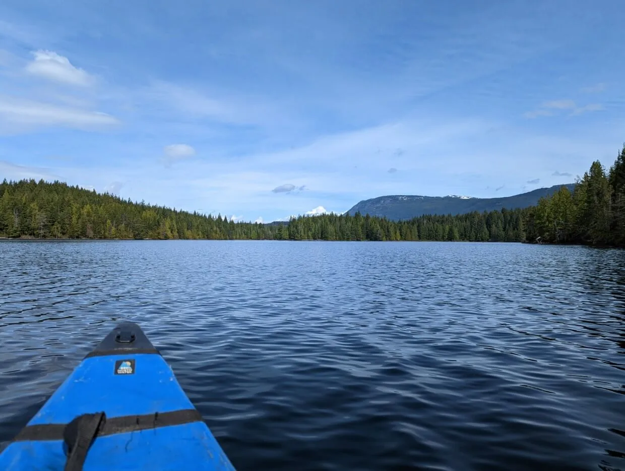 Canoe view of calm lake, which is lined with trees. A forested hill is visible in the background