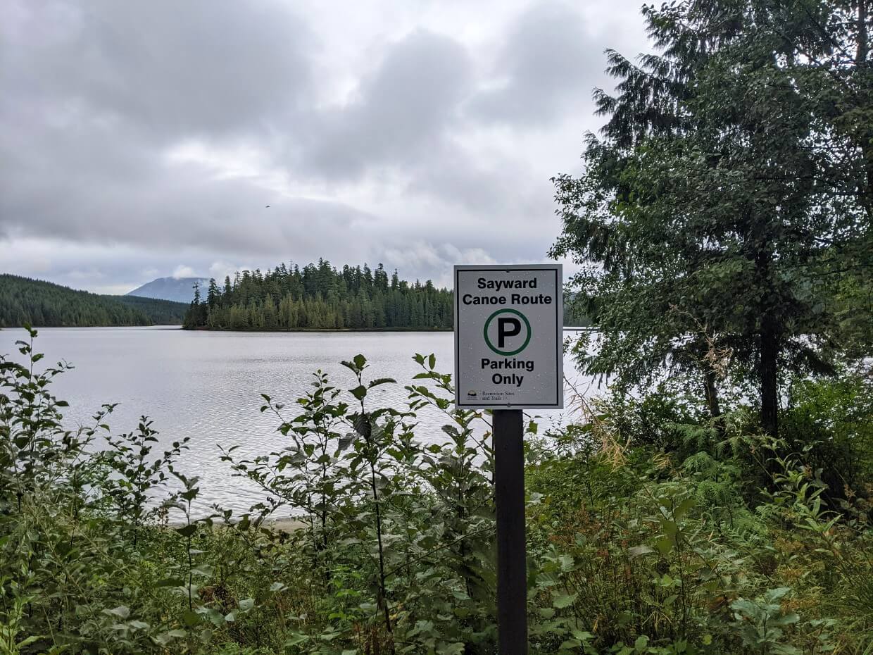 Sign for Sayward Canoe Route parking at Pacific Yew Recreation Site. Just beyond the sign is low lying brush and a lake.