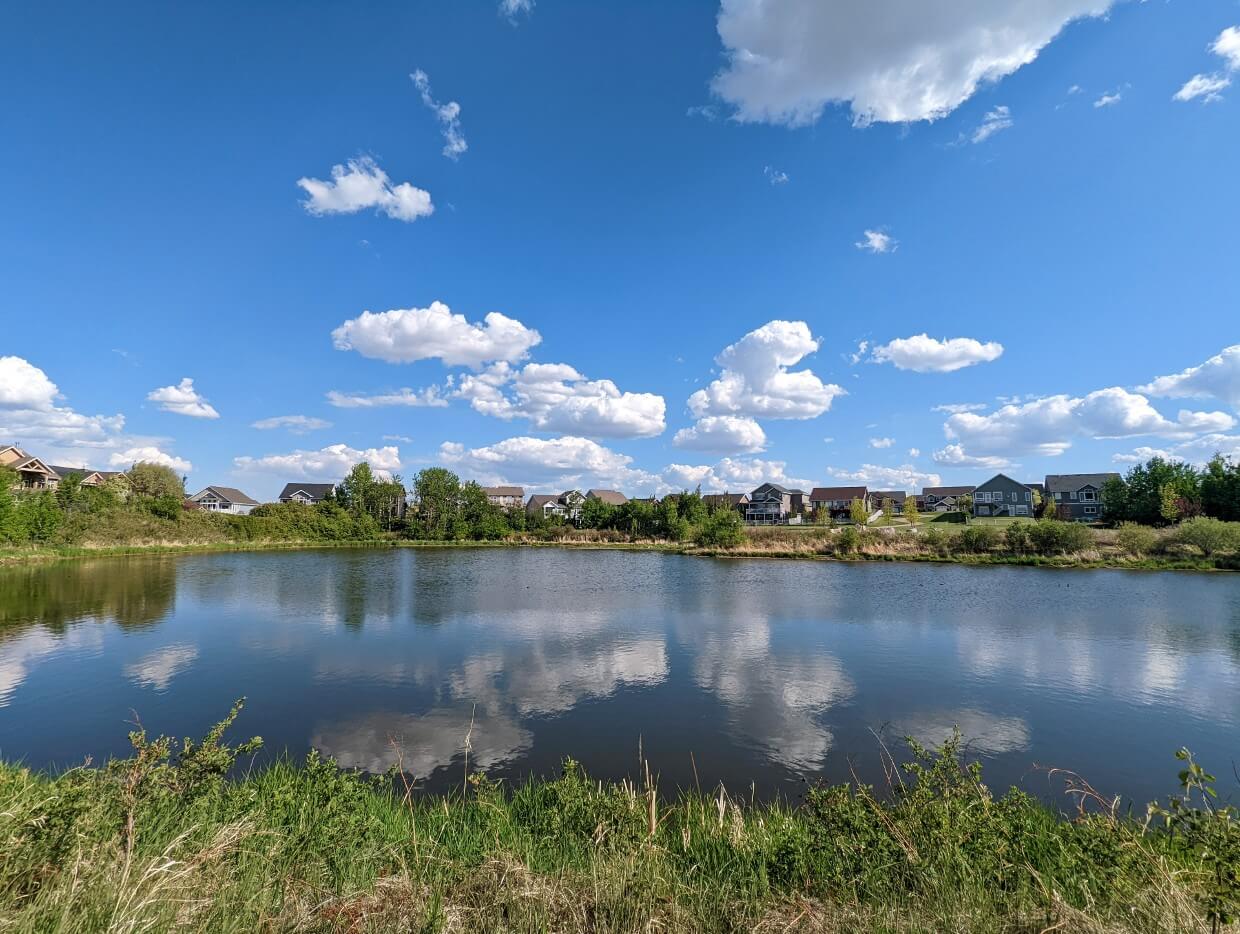 Calm reflective lake in Lacombe with blue skies and clouds, houses visible in background