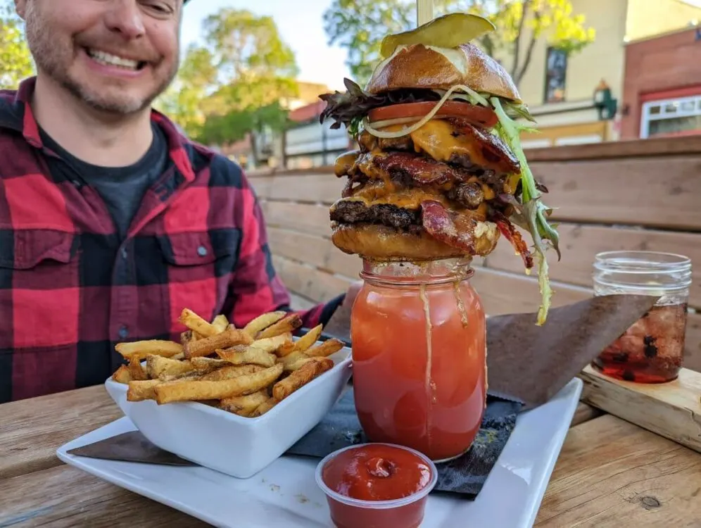 A huge multi-layered bacon burger sits on top of a Caesar cocktail, with a bowl of fries on the side. JR is visible in the frame and is smiling while looking down at the meal