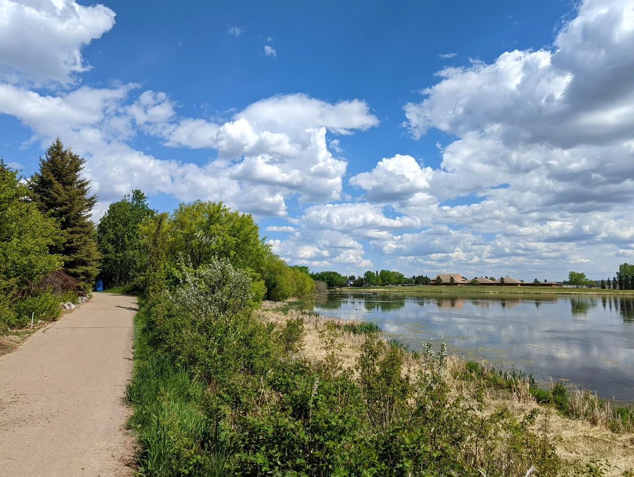 Paved trail on left runs along shoreline of calm reflective lake in Lacombe