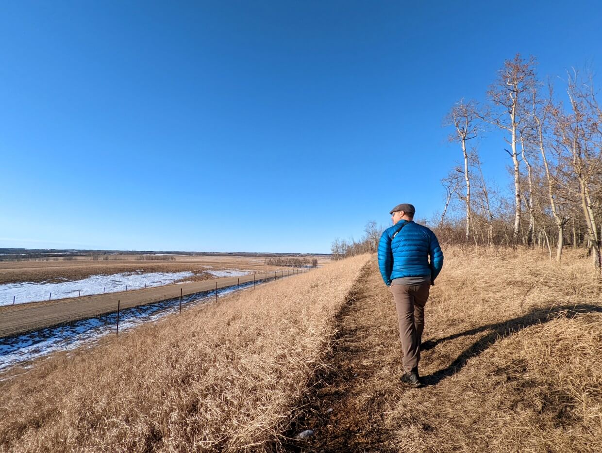 Back view of JR hiking on dry trail in late winter in Kuhnen Park, Lacombe, looking out to sweeping views of prairie