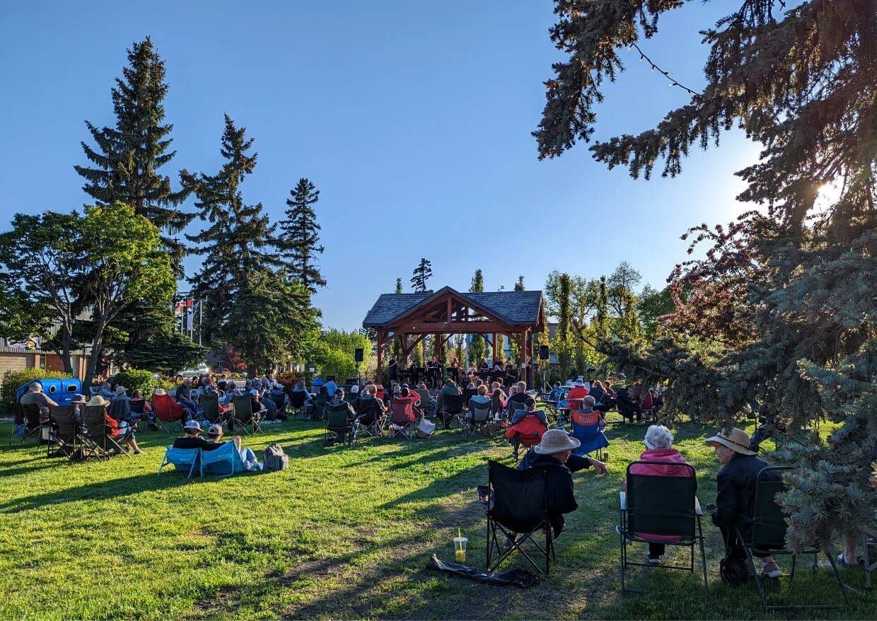 Back view of Music in the Park concert, with many people sat on grass and on chairs, watching music on stage