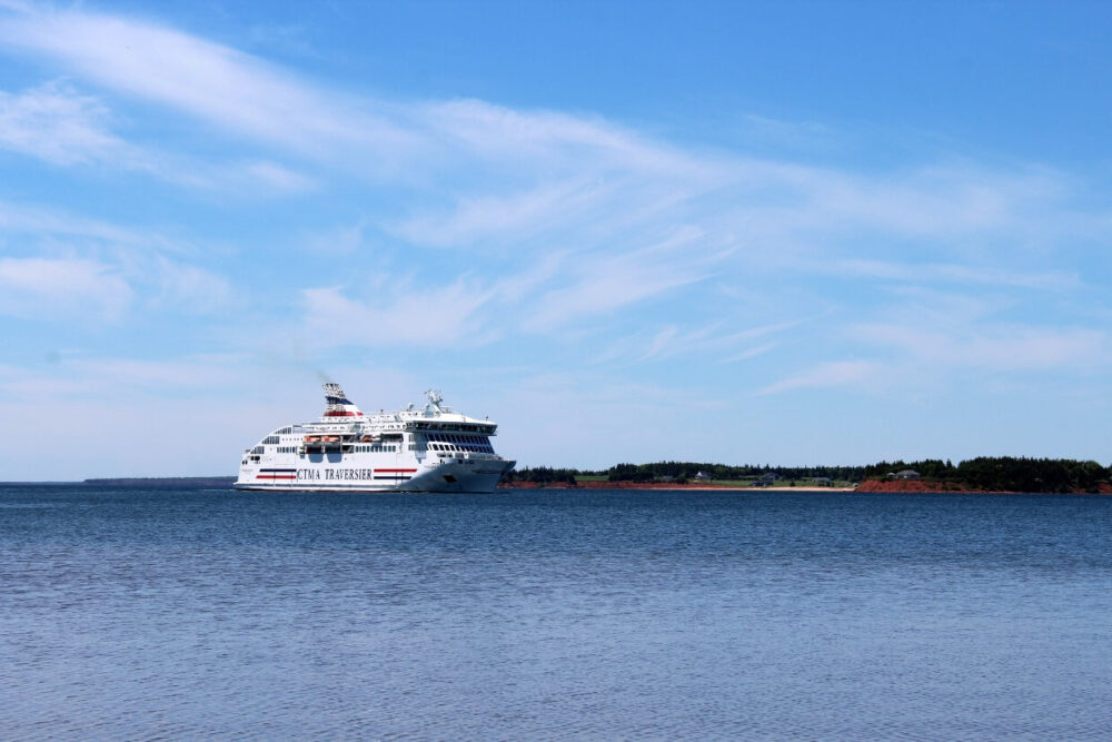Large body of water with the CTMA ferry arriving into Souris on the distant shore. The water is rippling from the boat and the sky is blue with wispy clouds.