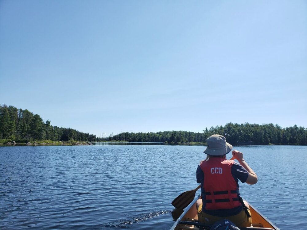 A woman is paddling on a lake. The waters are calm and the blue sky overhead is clear. In the distance the lake is framed with trees. 