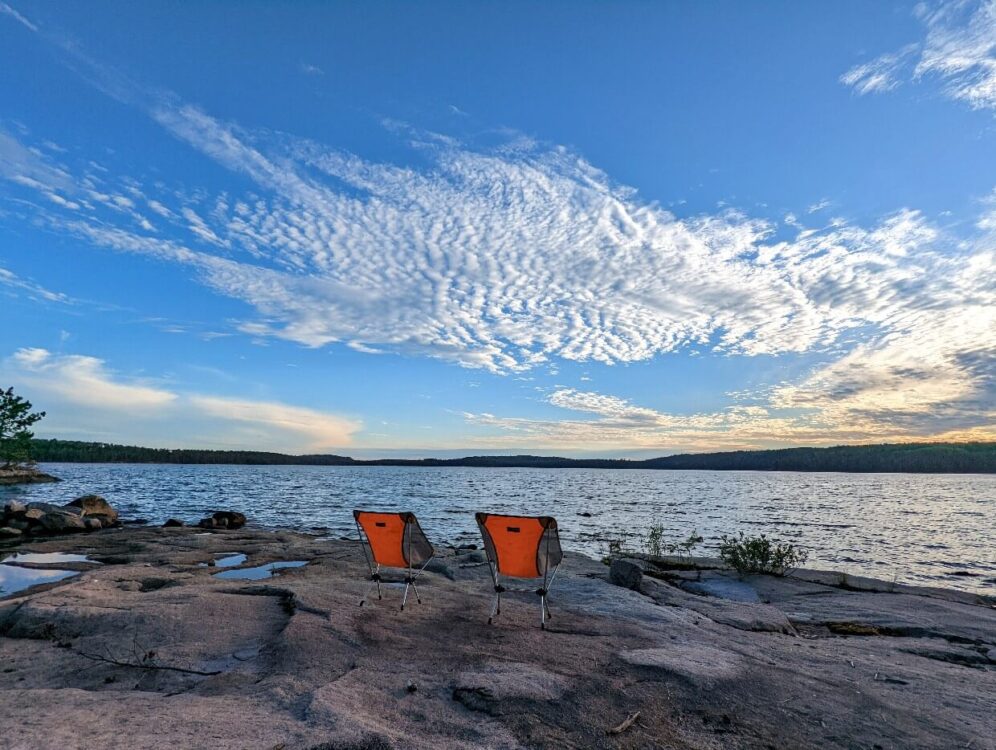 Two folding camp chairs sit on a rocky shoreline looking out onto Jean Lake. Surrounding the large is dense forest on all sides. The sky is a bright blue with wispy clouds stretching out into the distance. 