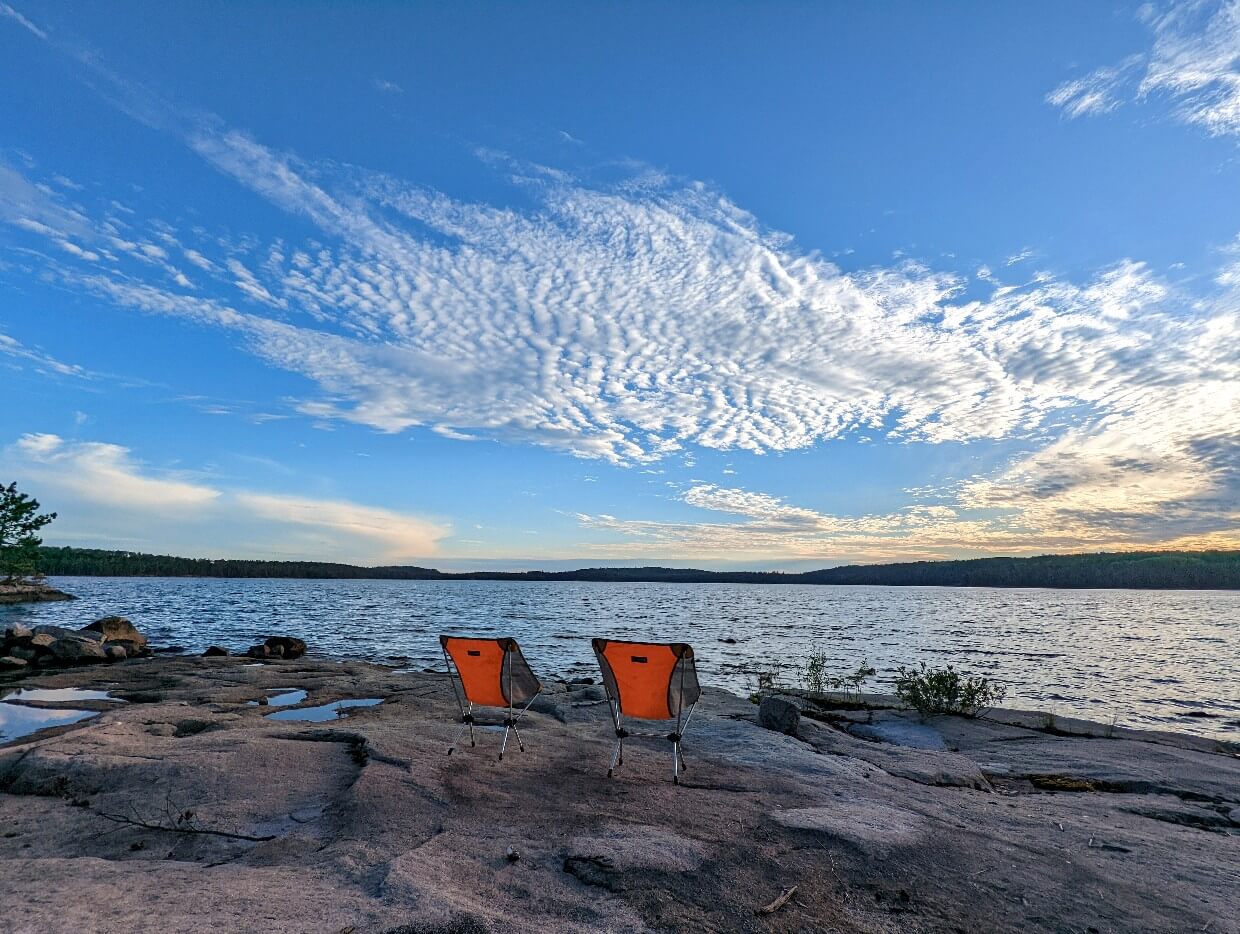Two folding camp chairs sit on a rocky shoreline looking out onto Jean Lake. Surrounding the large is dense forest on all sides. The sky is a bright blue with wispy clouds stretching out into the distance.