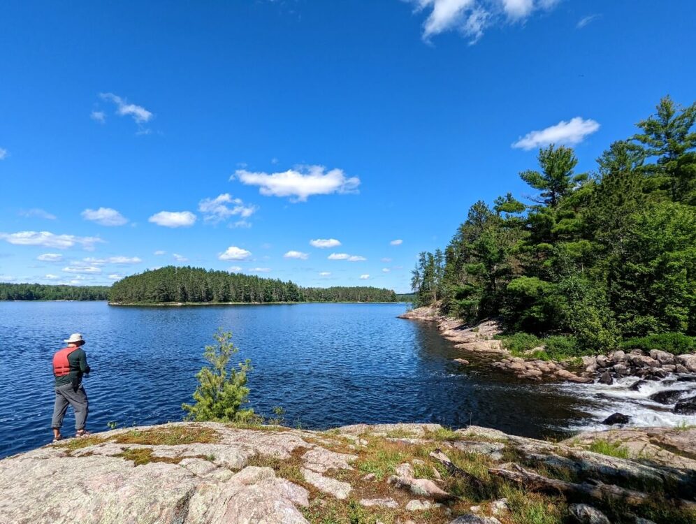 A man standing to the left of the frame is fishing from the rocky shore of Quetico Lake. To the right is a small rapids. The lake is wrapped with rocky shores and dense forests. Above the sky is a bright blue with some small white fluffy clouds.