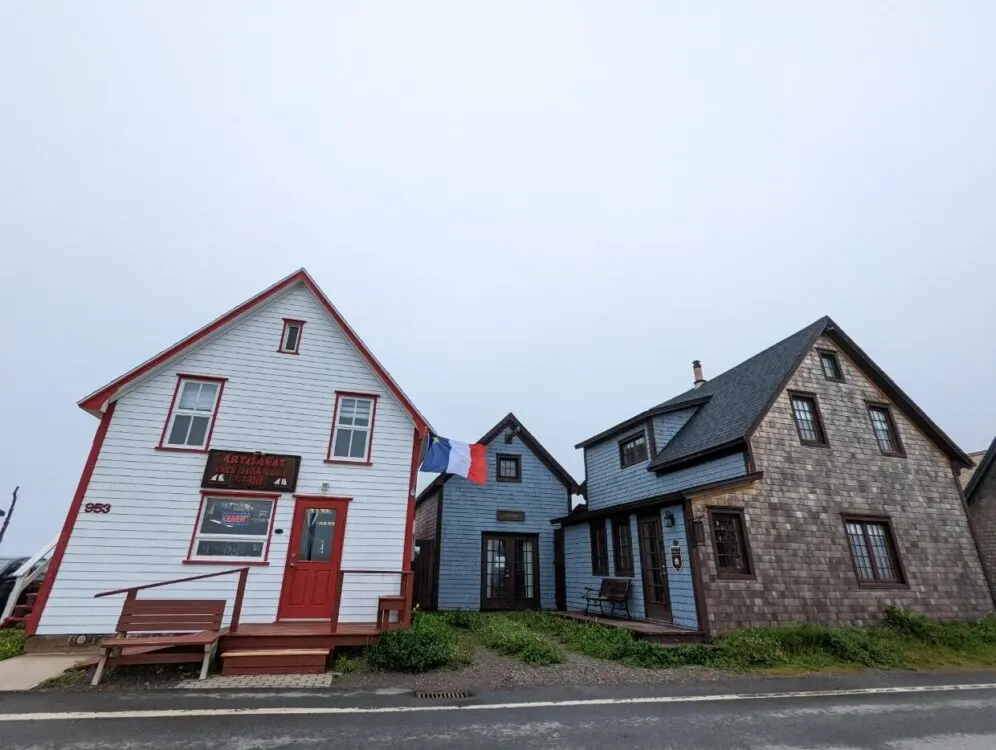 Historical wooden buildings at La Grave, with white painted building on left and blue two story building on right. There is an Acadian flag in the middle