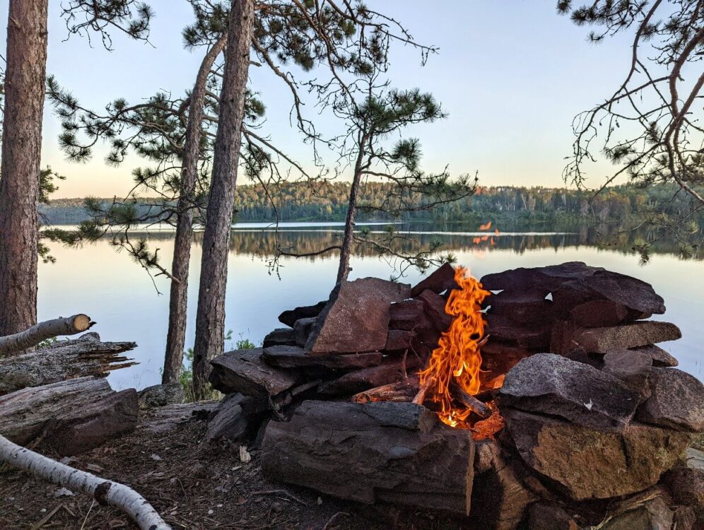 A campfire blazes in a fire pit made up of stacked stones. The fire sits on the edge of McAlpine Lake. Behind the fire is a view of the lake and forest reflecting in it. The sky is clear, with warm tones of a sunset fading.
