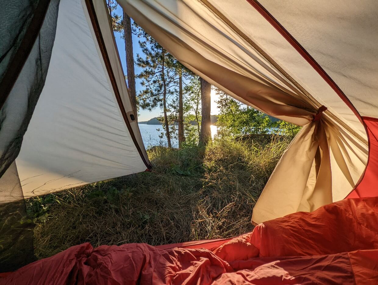 Looking out through a tent onto the shores of McAlpine Lake. In the forefront is disturbed grass. In the distance the lake can be seen peeking through the few trees, along with the sun and clear blue sky.