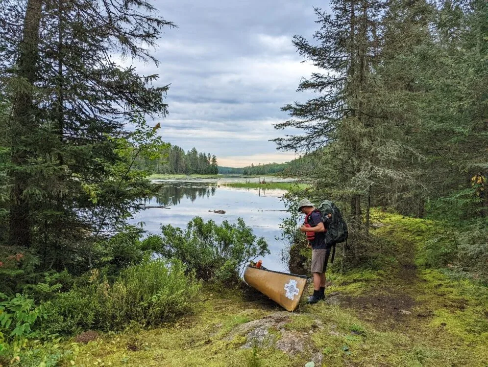 A man stands with a large backpack beside a canoe. The canoe is on the edge of a lake that is surrounded by dense forest. In the distance the lake is swampy and the sky is full of clouds.
