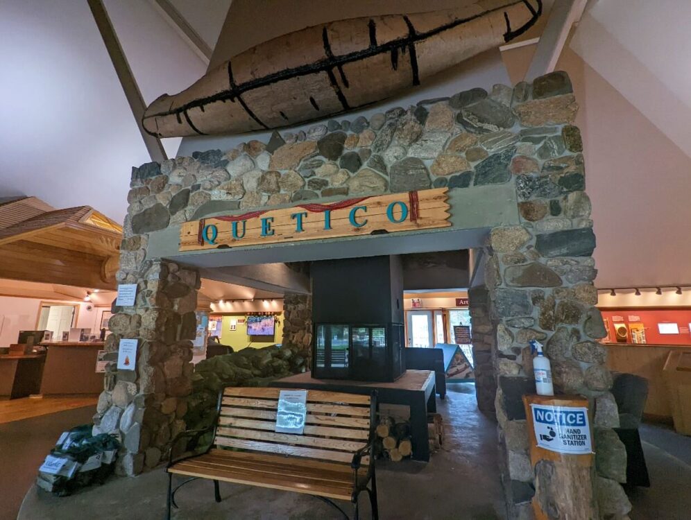 Inside the Dawson Trail Heritage Pavilion sits a massive stone gate with a wooden sign saying Quetico. Above the gate an old birch bark canoe hangs on the wall and in the entrance is a bench.