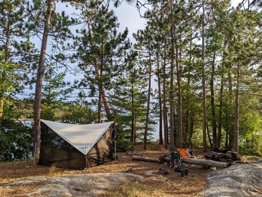 A tent and campsite set up on Quetico Lake. The tent is sitting on grass but parts of the campsite are rocky. The lake can be seen in the distance through the line of trees.