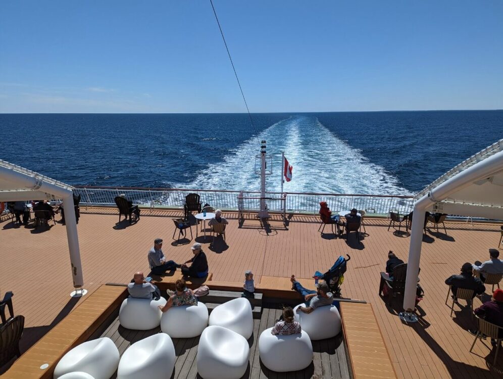 The aft deck of a ferry with people seated in chairs scattered around the deck. Off the deck the wave from the ferry can be seen in the water under a clear blue day.