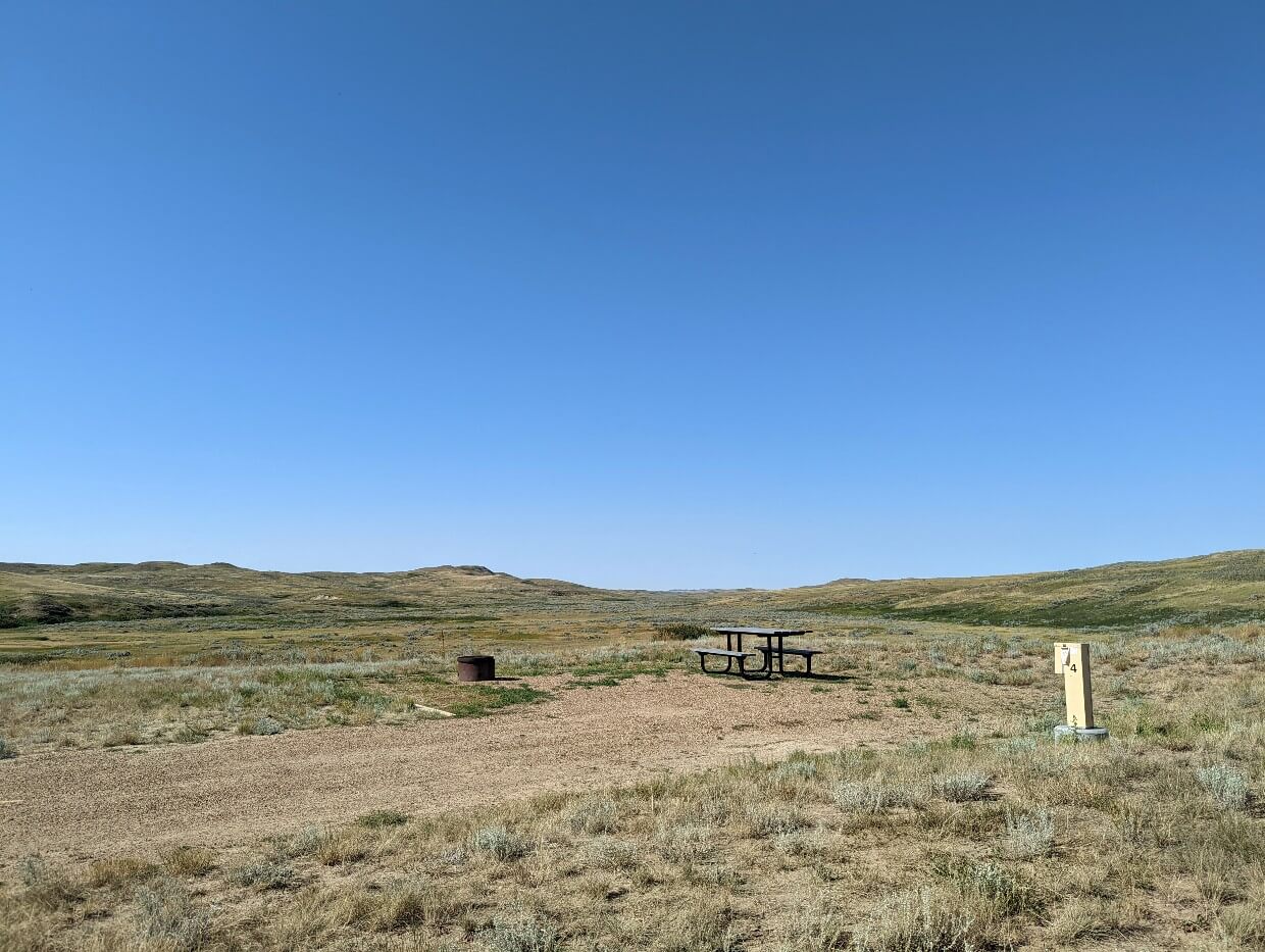 Campsite at Rock Creek Campground, an open dirt area in front of rolling hill scenery with picnic table and fire pit