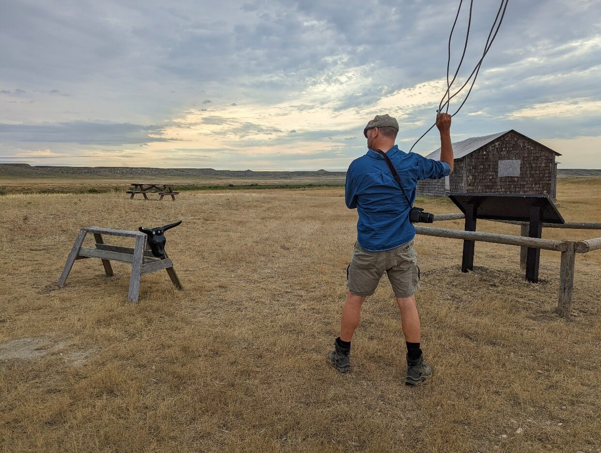 Back view of JR lassoing a practice cow in Grasslands National Park, with ranch building behind