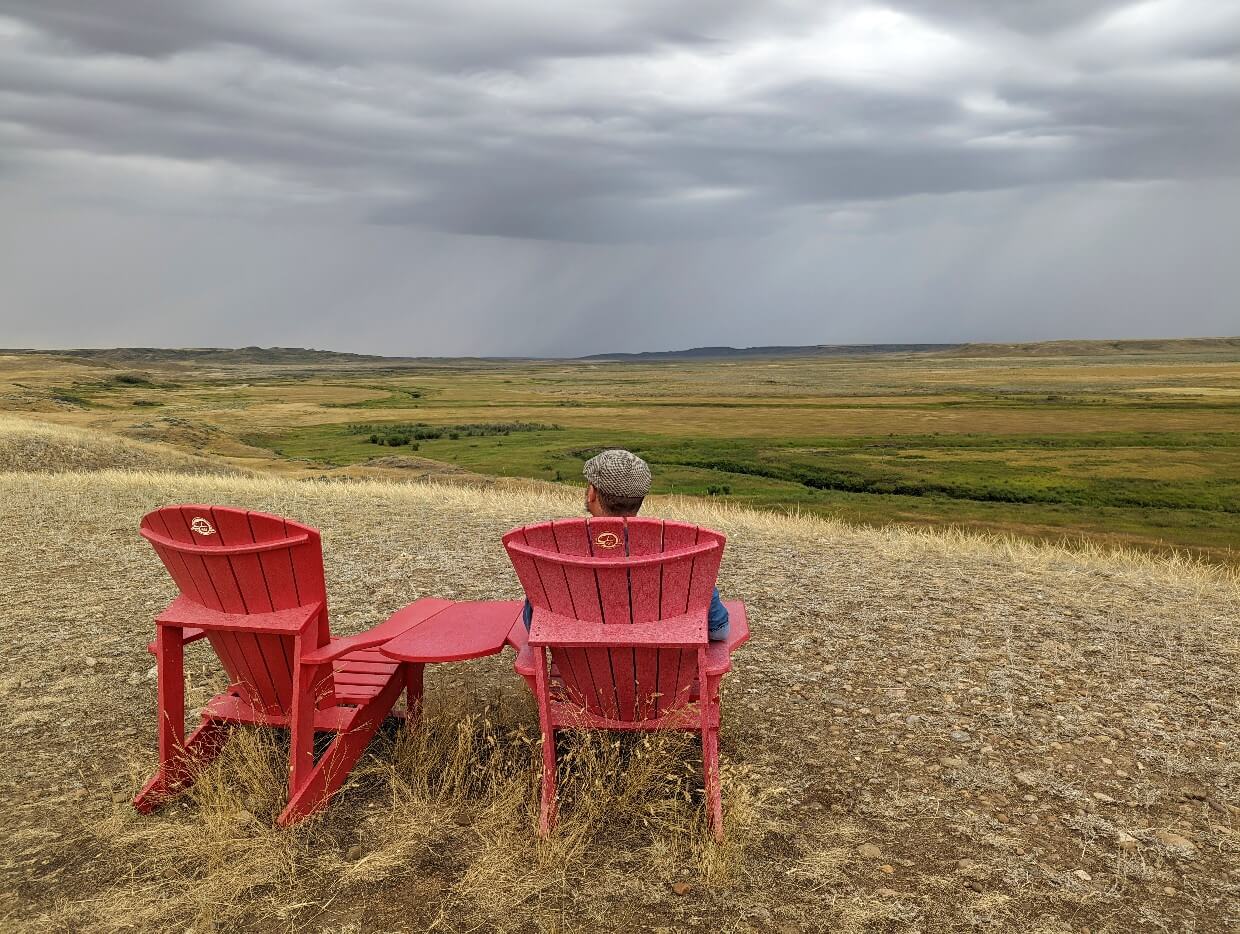 Back view of JR siting in red Parks Canada Adirondack chair in front of rolling prairie landscape in Grasslands National Park's West Block