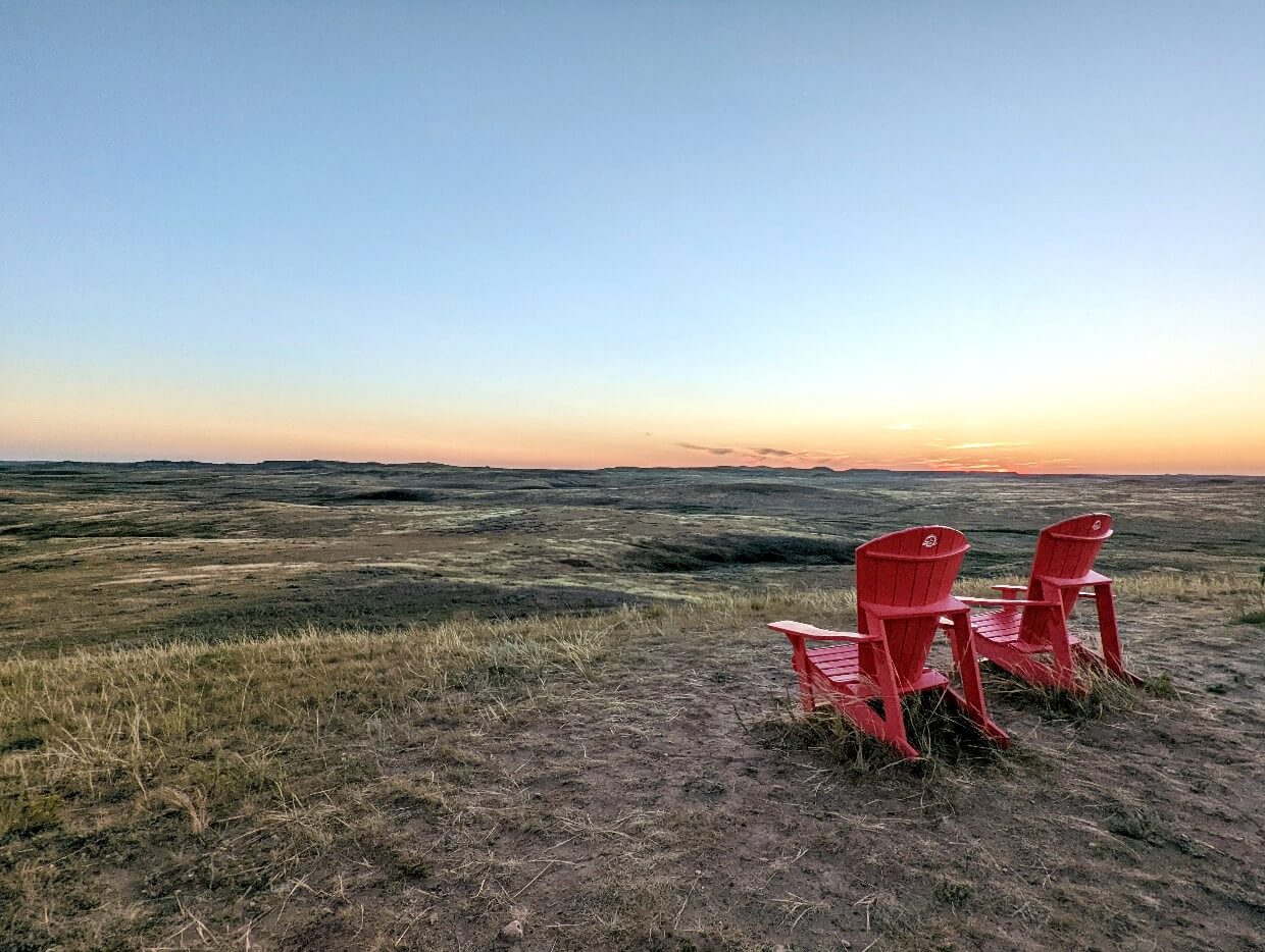 Back view of red Parks Canada Adirondack chairs in front of sunset over grasslands scenery