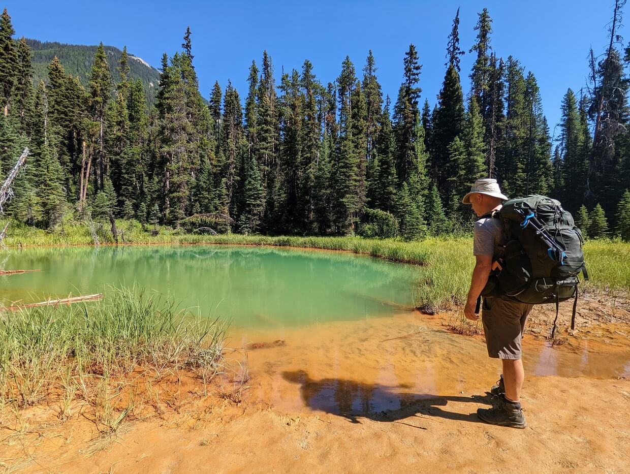 Back view of JR standing next to green coloured pool, with rich ochre mud surrounding it