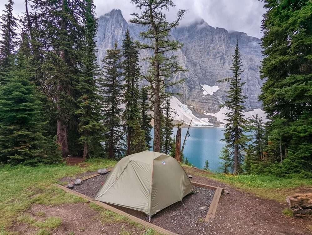 Set up tent on gravel pad in front of turquoise coloured Floe Lake, backdropped by soaring mountains