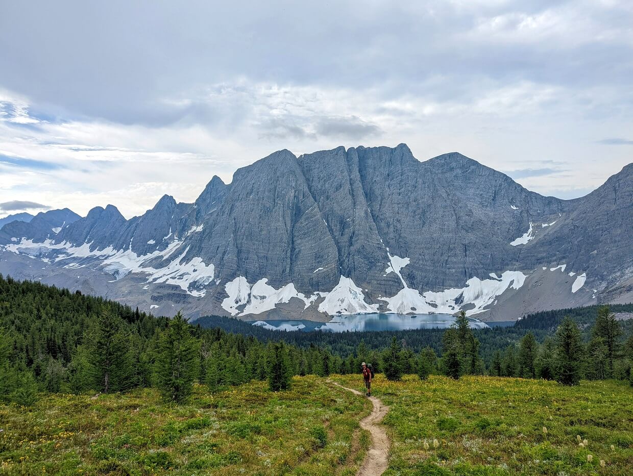 Back view of JR hiking down trail through wildflower meadows, with turquoise Floe Lake visible in background with soaring mountains as a backdrop