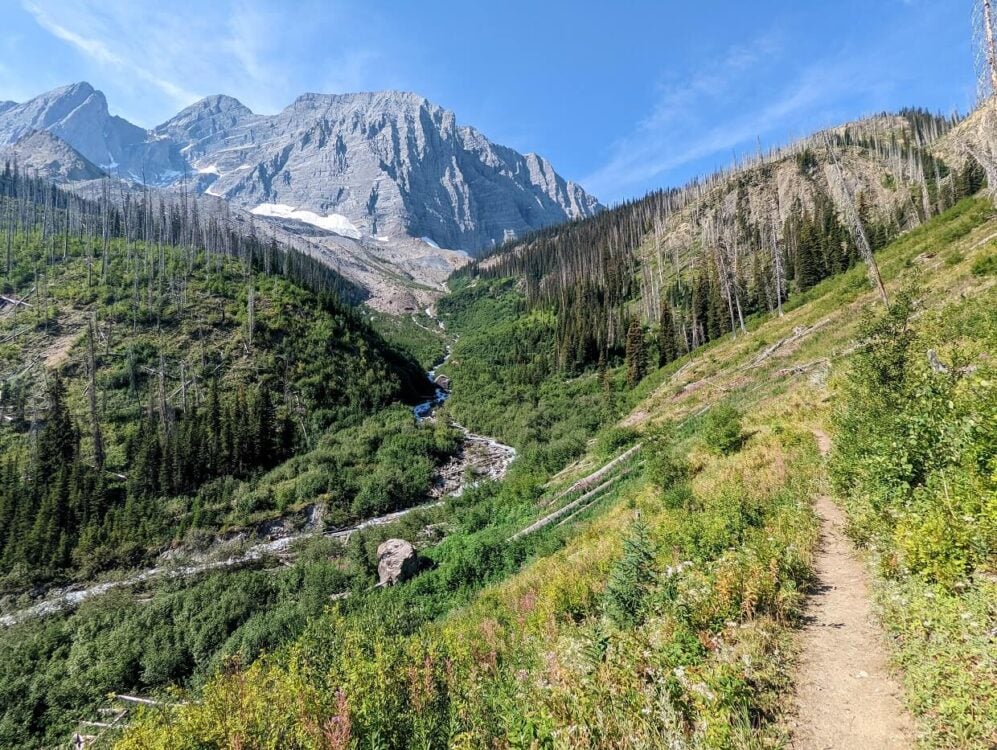 A small dirt trail leads away from camera towards mountain range. There is a river winding its way down from the mountain to the left