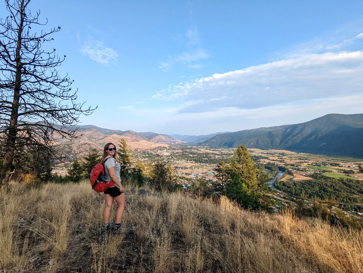 Side view of Gemma with red backpack looking at camera at top of Saddle Mountain. Grand Forks is visible below, surrounded by golden scenery