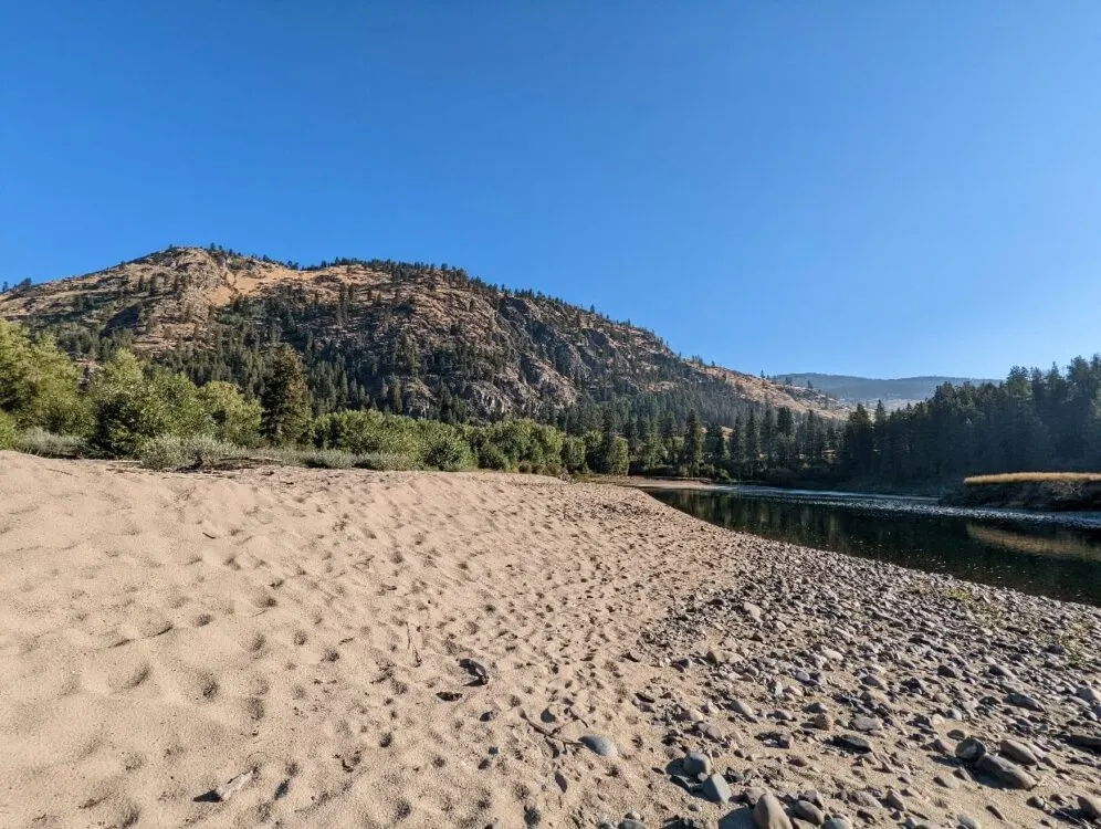 Looking across sandy beach to calm river in Boothman's Oxbow Provincial Park which is backdropped by forested mountain