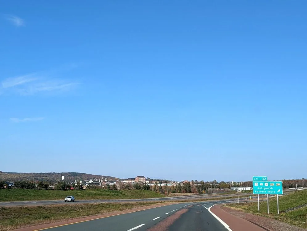 Vehicle view looking towards highway and across to town of Antigonish. There is a green road sign on the right stating 'Antigonish, Eastern Shore' with an arrow