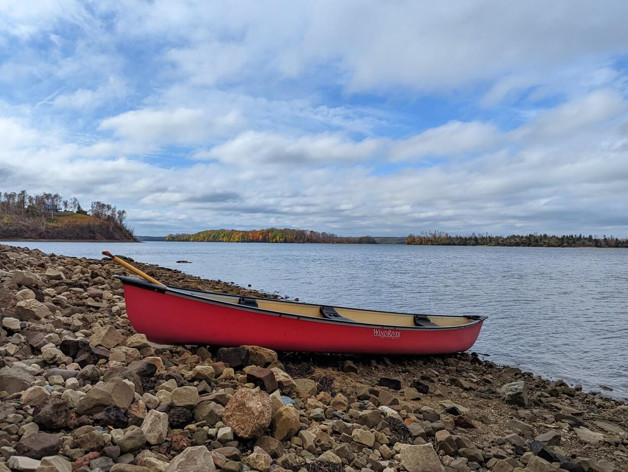 Red canoe on shore at Fossil Farms with calm ocean in background, some autumn colours visible in trees on other side of bay