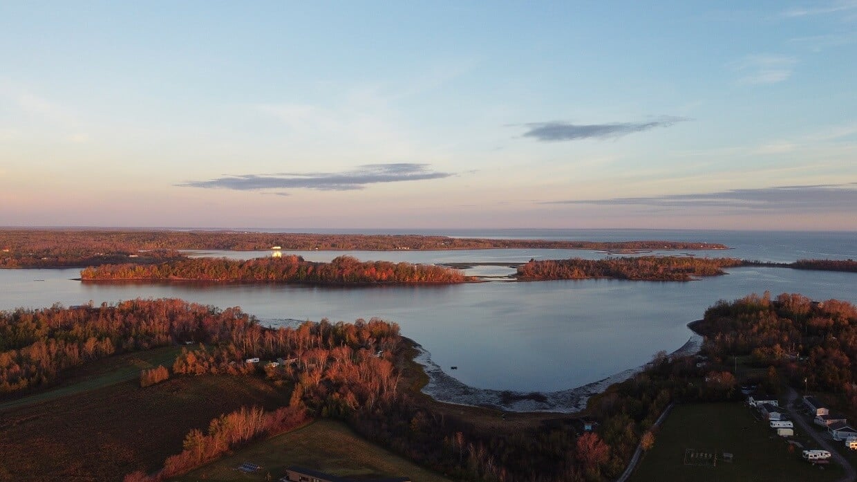 Drone photo of Fossil Farms, with cottages on left, beach in middle and island (with white church building) behind. Sunset colours with some clouds in the sky