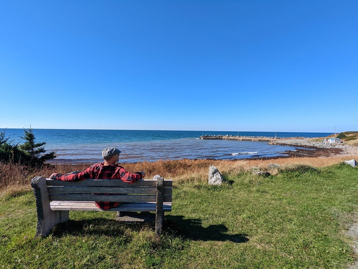 Back view of JR sat on bench at Livingstone's Cove Wharf Park on Cape George Scenic Drive, looking out to rock beach