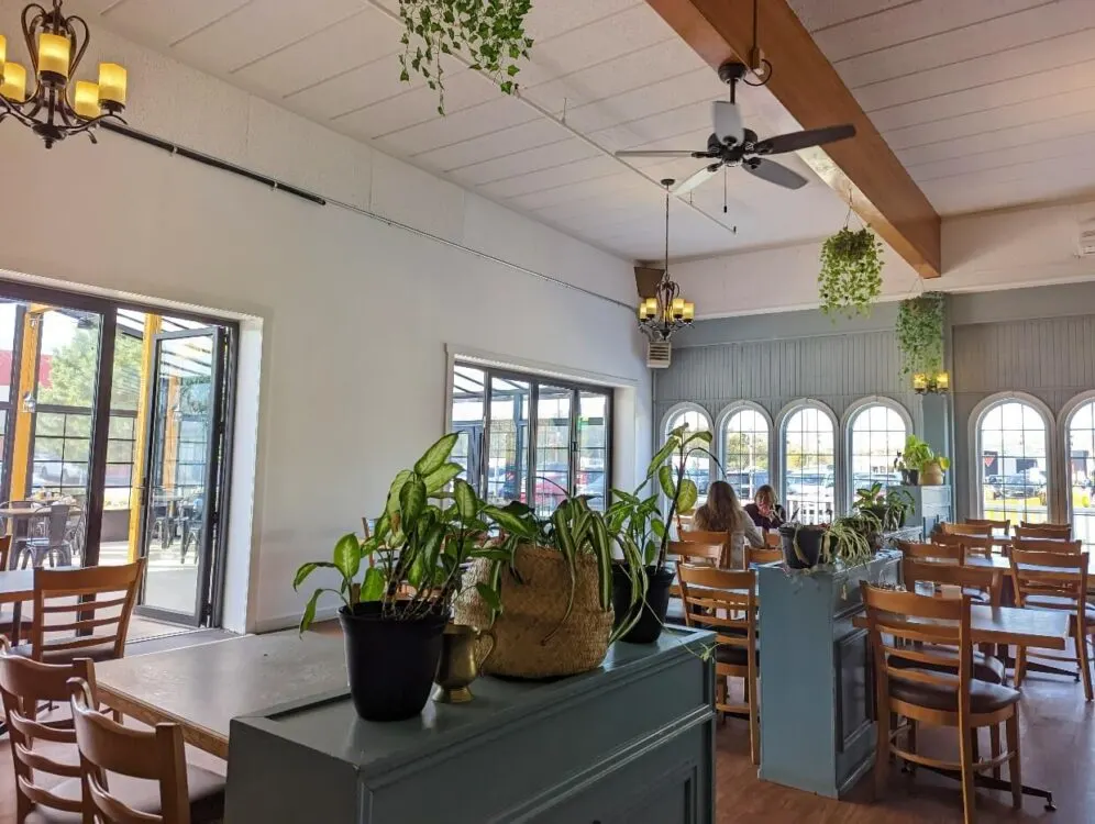 Looking across bright and airy dining space at Justamere Cafe and Bakery, with high ceilings, many plants, wooden tables, wooden floor and white walls. One table is occupied