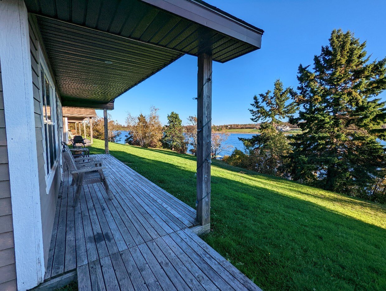 Side view of Pomquet Beach Cottages with covered wooden deck looking out to sloping lawn and ocean views, with scattered trees