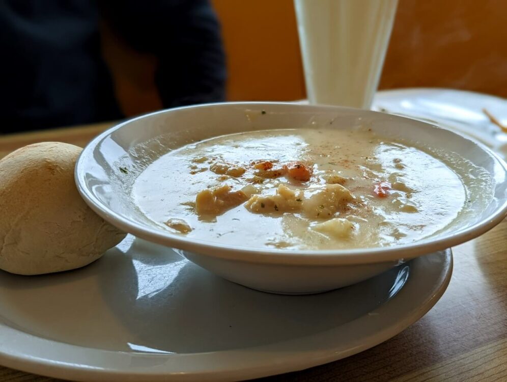 Close up of seafood chowder dish at Snow Queen restaurant with large bowl filled with creamy liquid and pieces of seafood. There is a bread roll next to the bowl