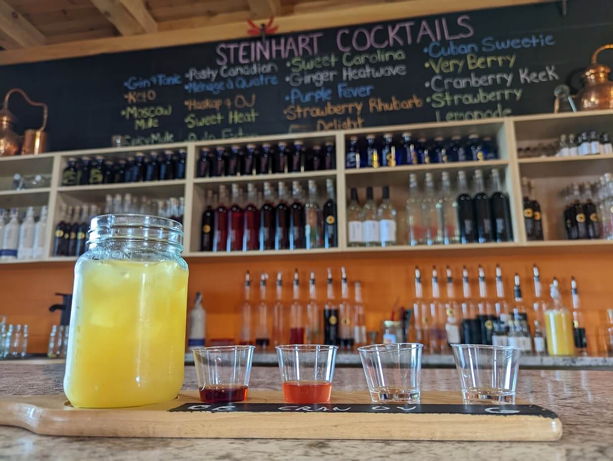 Close up of flight of spirits at Steinhart Distillery, with cocktail on left. Flight is in front of chalkboard and bar