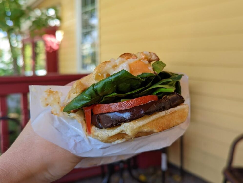 Close up of waffle sandwich held in hand, featuring eggplant, tomato and spinach