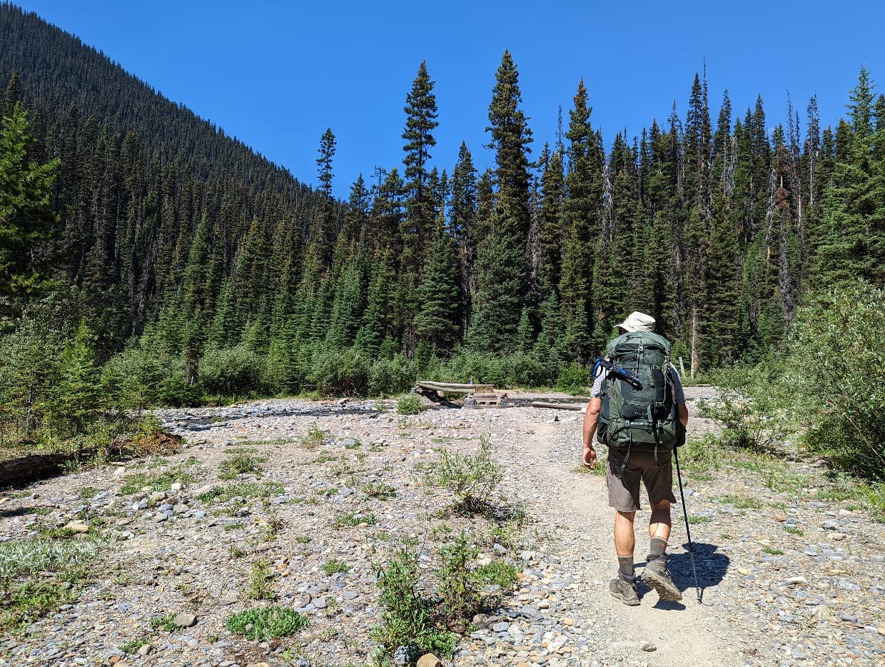 Back view of JR hiking towards river with bridge at Helmet Ochre Junction campground on Rockwall Trail