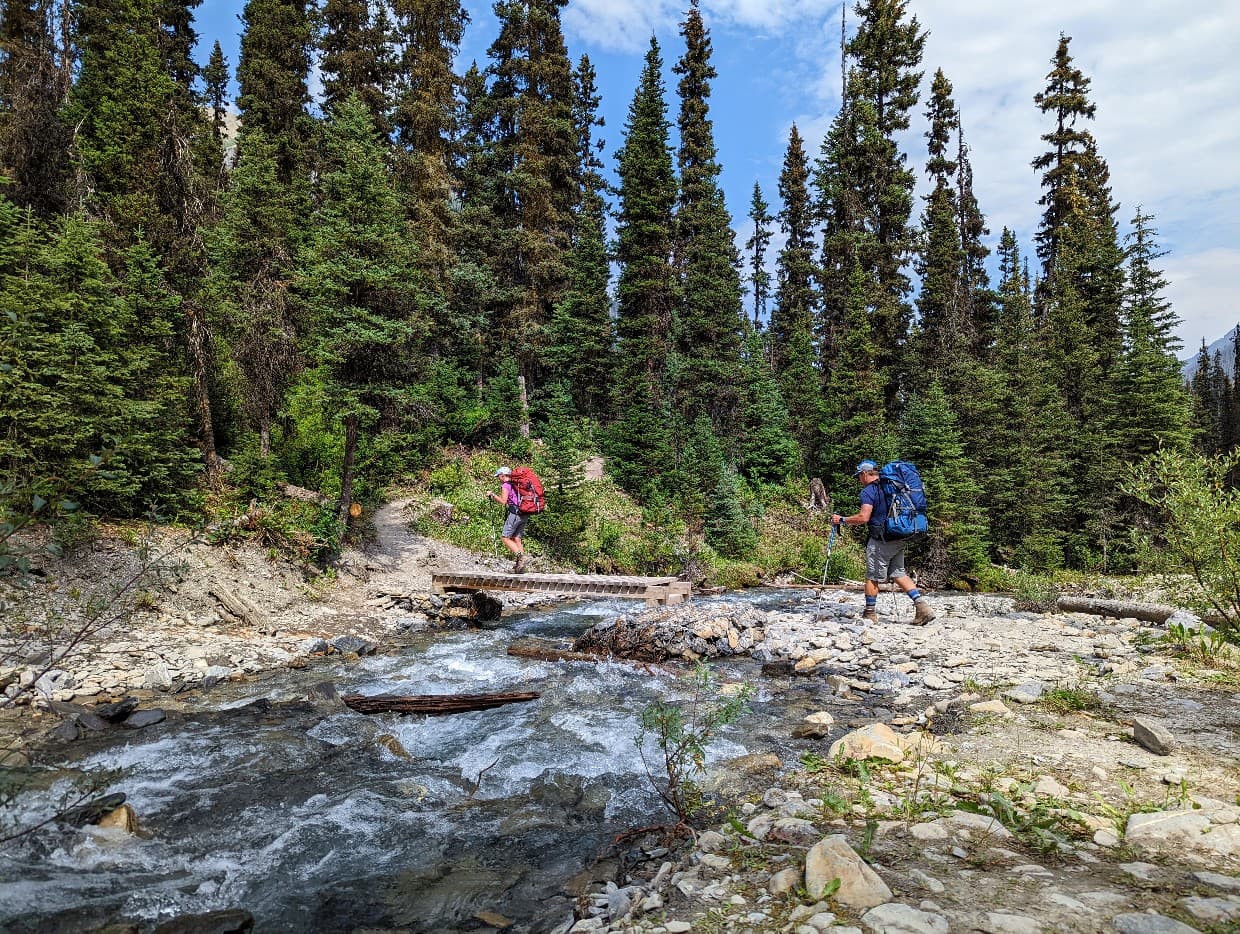 Two hikers with colourful backpacks cross a wooden bridge over a fast flowing creek