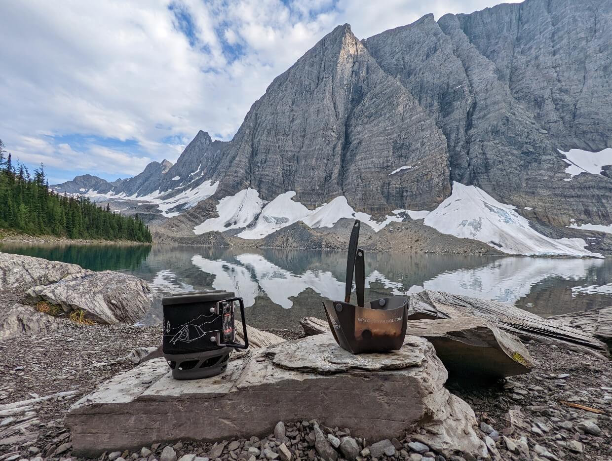 Cooking pot and bowl with food on rock in front of reflective Floe Lake, with towering mountains behind