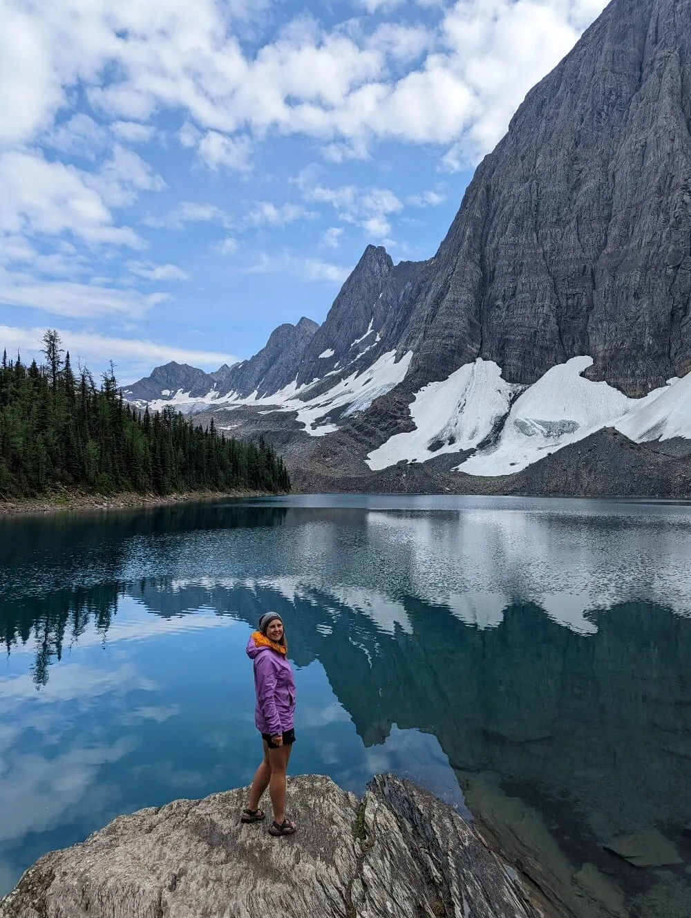 Gemma stands on rock in front of Floe Lake, looking at camera. The calm surface of Floe Lake reflects the huge mountains above it