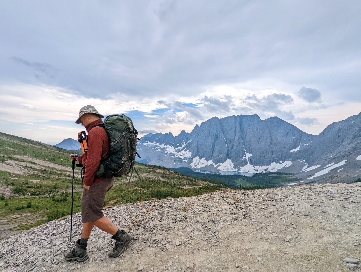 Side view of JR hiking down scree section of Rockwall Trail, wearing large backpack and bear spray holster, with Floe Lake in background